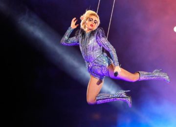 Lady Gaga in costume argentato, appesa a dei cavi, simula un volo divino illuminata di viola