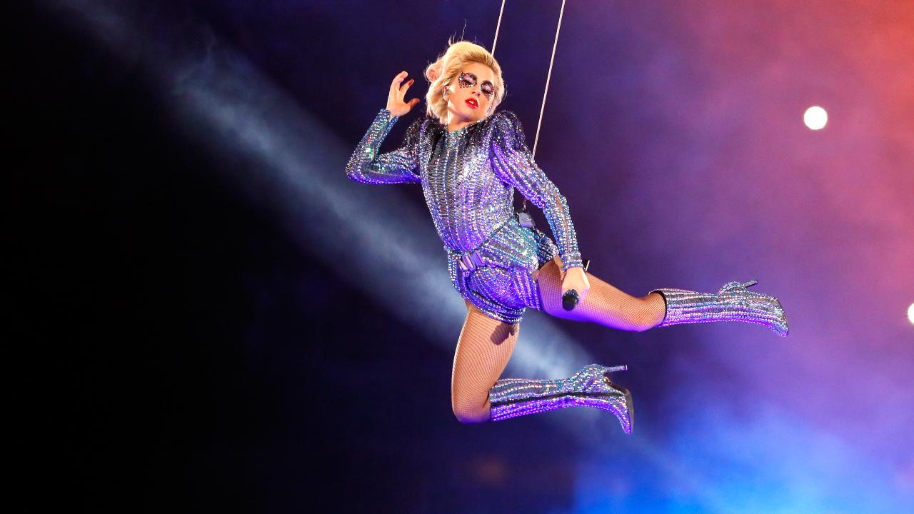 Lady Gaga in costume argentato, appesa a dei cavi, simula un volo divino illuminata di viola